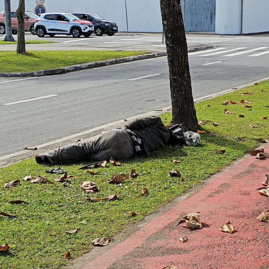 Pessoa em situação de rua deitada na grama, encostada em uma árvore, próxima a uma ciclovia e a uma avenida em Florianópolis. Ao fundo, há carros parados no semáforo.