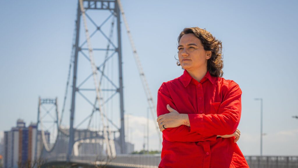 Foto da Carla Ayres em frente à Ponte Hercílio Luz,  que representa a conexão entre a ilha e o continente.